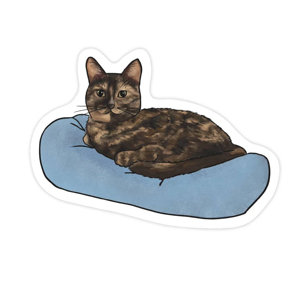 Sticker of a tortoiseshell cat lying on a blue pillow with a white border.